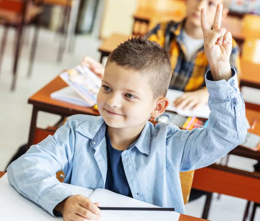 A young male student raises his hand in the shape of a V to at his desk in a classroom.