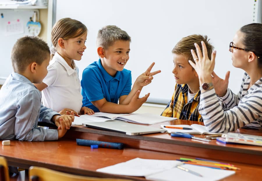 Three students gather around a female teacher's desk to ask questions and talk about their lesson.