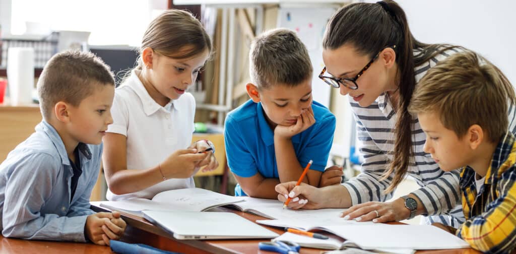 A female teacher with four students gather around her desk to review a workbook.